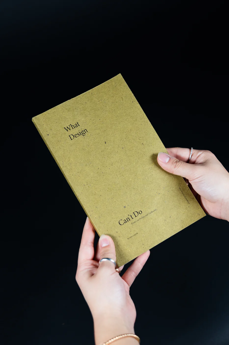 Hands holding the finished book, an olive speckled cover with What Design Can’t Do set in a light serif