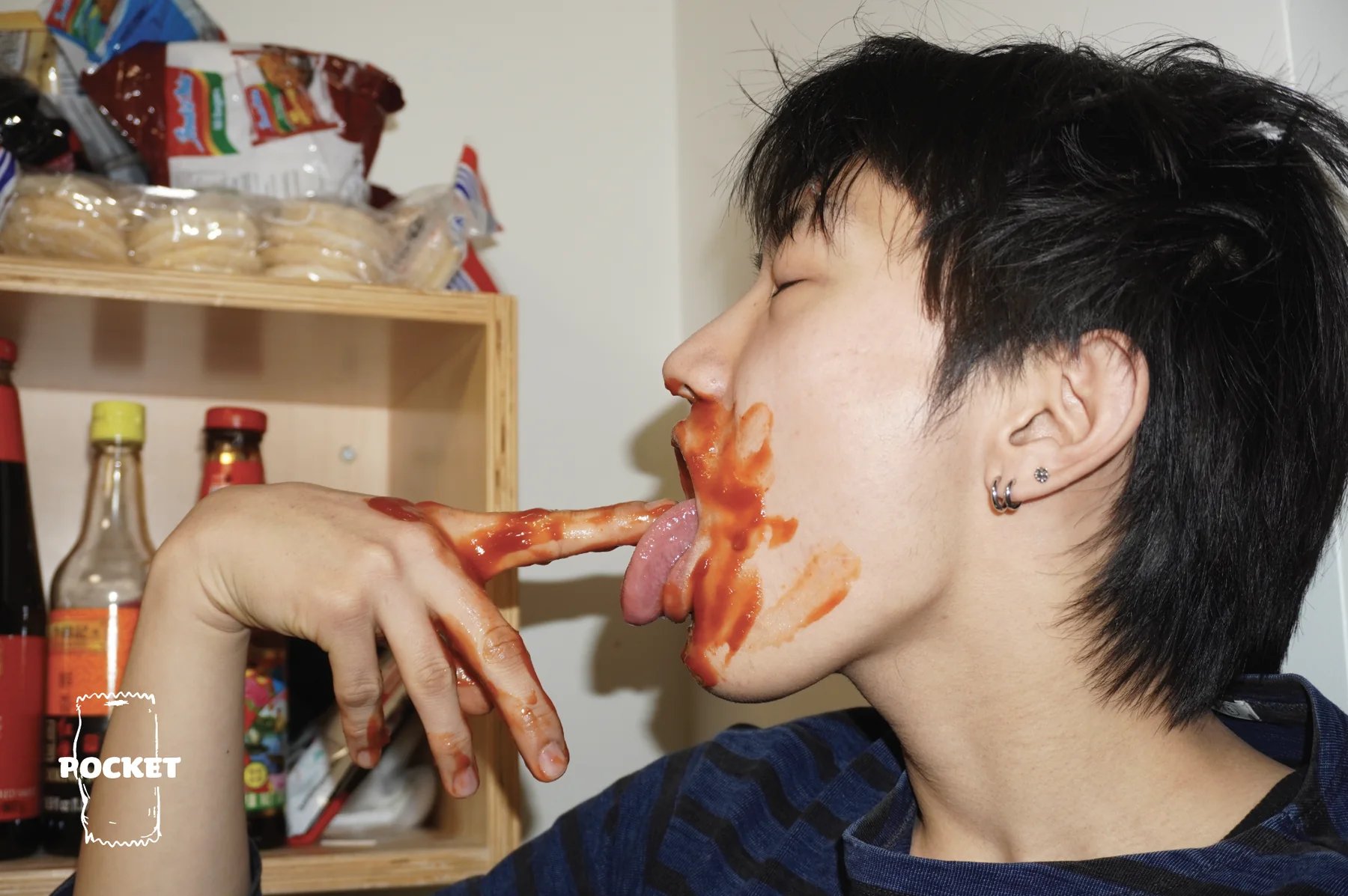 A model with sauce on their face and fingers in front of a shelf of sauces, with the Pocket Sauce packet logo